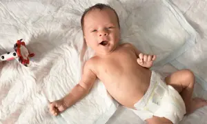 Ein fröhliches Baby, etwa 6 Wochen alt, liegt auf einer weißen Unterlage und blickt neugierig in die Kamera. Neben ihm liegt ein buntes Spielzeug.
