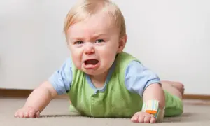 Ein weinender **baby 16 wochen** alt liegt auf dem Bauch auf einem Teppich. Es hat blonde Haare und trägt ein grünes Oberteil über einem blauen Hemd.