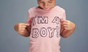Ein Kind mit Mütze zeigt stolz auf sein rosa T-Shirt mit der Aufschrift "I'M A BOY!". Ein Bild, das die Identität von transgender Kindern feiert.