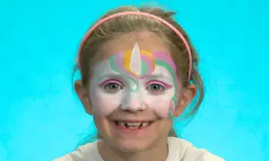 Ein Mädchen mit einem bunten Einhorn-Schminken für Kinder. Das Gesicht ist weiß grundiert, mit einem weißen Horn und bunten Bögen in Rosa, Gelb und Grün.