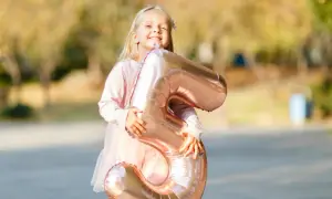 Ein glückliches, blondes Kind von 5 Jahren hält stolz einen großen, roségoldenen Ballon in Form der Zahl 5.
