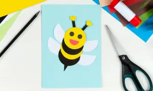 Süße Biene basteln: Gelber Kopf mit Herzfühler, schwarze Streifen, weiße Flügel auf hellblauem Papier. Schere und Klebestift liegen bereit.