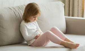Ein Kleinkind mit blonden Haaren sitzt auf einer Couch und hält sich den Bauch. Es scheint, als hätte das Kleinkind Erbrechen ohne weitere Symptome.