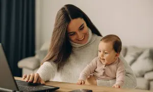 Lächelnde Mutter mit Baby auf dem Schoß am Laptop. Ein Bild, das den Spagat zwischen Beruf und Familie zeigt, oft verbunden mit dem Gefühl von Mama Burnout.