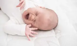 Ein Baby mit weit geöffnetem Mund und geschlossenen Augen, das sich im unruhigen Schlaf windet. Seine kleinen Hände sind nach oben gestreckt.