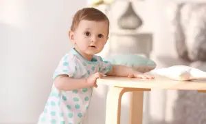 Ein neugieriges Baby, etwa 1 Jahr alt, stützt sich auf einen Holztisch. Es trägt ein weißes T-Shirt mit blauen Tupfen.