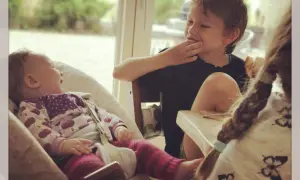 Drei Kinder am Tisch: Ein Baby lacht, ein Junge isst, ein drittes Kind mit Zopf ist im Vordergrund.