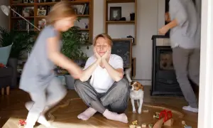 Chaos im Wohnzimmer: Kinder rennen herum, ein Hund schaut neugierig, und die mama steht kopf. Sie sitzt auf dem Boden, die Augen geschlossen, während das Leben um sie herum tobt.