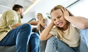 Ein Kind hält sich die Ohren zu, während Eltern im Hintergrund streiten. Das aggressive Verhalten der Erwachsenen belastet das Kind sichtlich.
