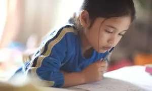 Ein **kind 6 jahre** alt, konzentriert sich auf seine Hausaufgaben. Es liegt auf dem Bauch und hält einen Stift in der Hand.