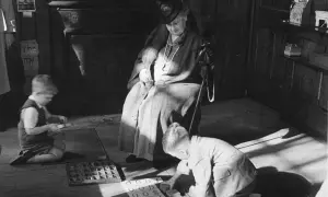 Maria Montessori beobachtet Kinder beim Lernen. Ein Junge kniet und sortiert Buchstaben, ein anderer übt mit Zahlen.