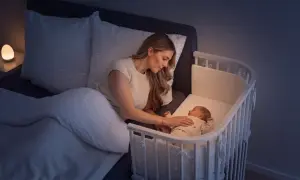 Mutter beruhigt ihr Baby im Beistellbett, um die Schlafregression zu überstehen. Sanftes Licht vom Nachttisch.