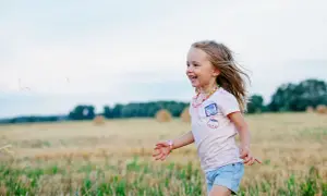 Ein fröhliches Kleinkind rennt lachend über ein Feld. Seine blonden Haare wehen im Wind, und es trägt ein buntes Halsband.