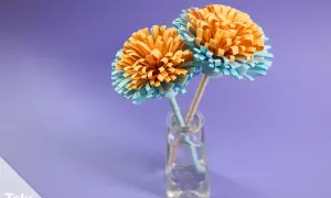Zwei selbstgebastelte Papierblumen in Orange und Hellblau stehen in einer kleinen Glasvase. Ein schönes Beispiel für blumenstrauß basteln.