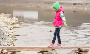 Ein Kind balanciert über ein provisorisches Holzbrett über einem Fluss. Das zeigt, was bedeutet selbstwirksamkeit: Mut, Vertrauen in die eigenen Fähigkeiten und das Überwinden von Hindernissen.