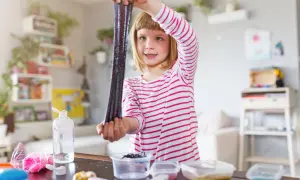 Ein Mädchen hält glitzernden, dunklen Schleim hoch, den sie gerade selber gemacht hat. Auf dem Tisch liegen Zutaten für weitere Schleim-Kreationen.