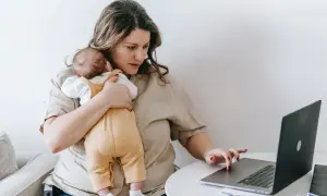 Alleinerziehend und im **burnout**: Eine Frau hält ihr Baby und arbeitet am Laptop.