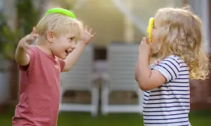 Zwei Kinder spielen im Garten. Der Junge mit einem grünen Ring auf dem Kopf lacht, das Mädchen hält ein gelbes Spielzeug. Ein Beispiel für gelebte sozialkompetenz definition.