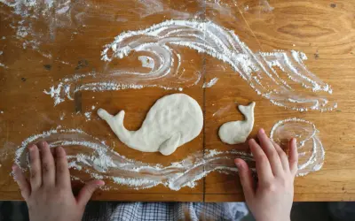 Kinderhände gestalten Salzteig-Ideen: zwei Wale schwimmen in Meereswellen aus Mehl auf einem Holztisch.