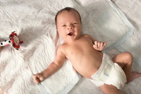 Ein fröhliches Baby, etwa 6 Wochen alt, liegt auf einer weißen Unterlage und blickt neugierig in die Kamera. Neben ihm liegt ein buntes Spielzeug.