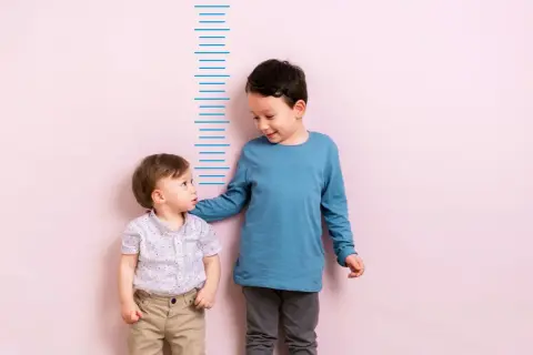 Zwei Jungen stehen vor einer Messlatte. Der ältere Junge, vielleicht 4 Jahre alt, legt dem kleineren seine Hand auf die Schulter.