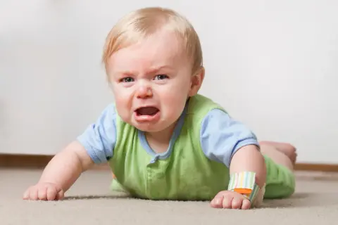 Ein weinender **baby 16 wochen** alt liegt auf dem Bauch auf einem Teppich. Es hat blonde Haare und trägt ein grünes Oberteil über einem blauen Hemd.