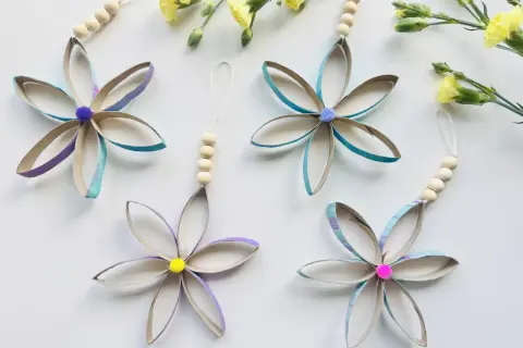 Bunte Blumen aus Klopapierrollen basteln: Vier hübsche Anhänger mit Holzperlen und Pompons, umgeben von gelben Blüten.
