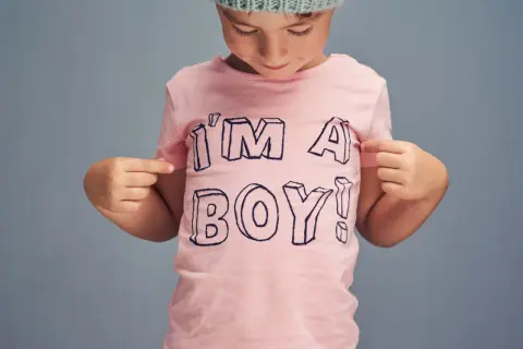 Ein Kind mit Mütze zeigt stolz auf sein rosa T-Shirt mit der Aufschrift "I'M A BOY!". Ein Bild, das die Identität von transgender Kindern feiert.