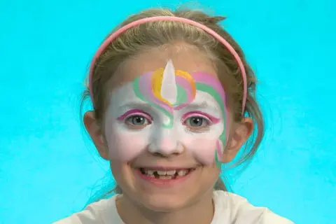 Ein Mädchen mit einem bunten Einhorn-Schminken für Kinder. Das Gesicht ist weiß grundiert, mit einem weißen Horn und bunten Bögen in Rosa, Gelb und Grün.