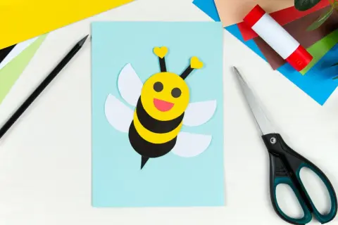 Süße Biene basteln: Gelber Kopf mit Herzfühler, schwarze Streifen, weiße Flügel auf hellblauem Papier. Schere und Klebestift liegen bereit.