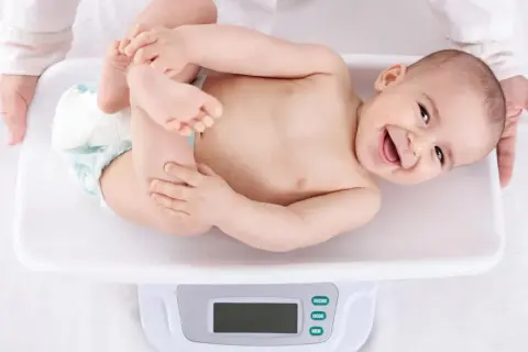 Ein lächelndes Baby liegt auf einer Waage für die U3 Untersuchung. Hände halten das Kind sanft.