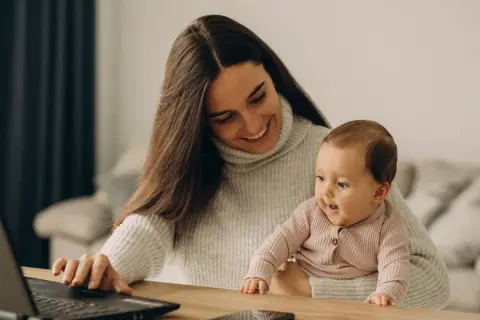Lächelnde Mutter mit Baby auf dem Schoß am Laptop. Ein Bild, das den Spagat zwischen Beruf und Familie zeigt, oft verbunden mit dem Gefühl von Mama Burnout.