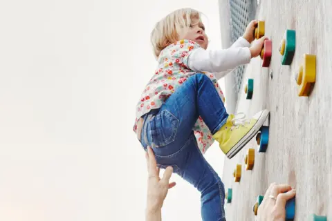 Kleines Kind klettert an einer Boulderwand, fördert seine Kompetenz. Hände helfen und sichern.