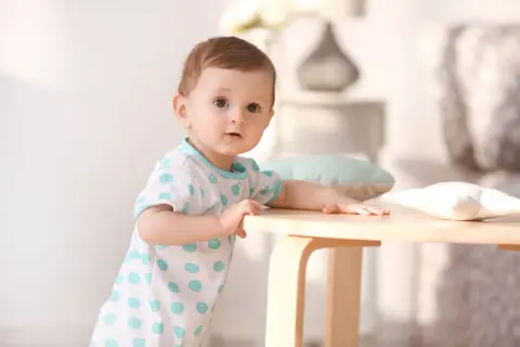 Ein neugieriges Baby, etwa 1 Jahr alt, stützt sich auf einen Holztisch. Es trägt ein weißes T-Shirt mit blauen Tupfen.