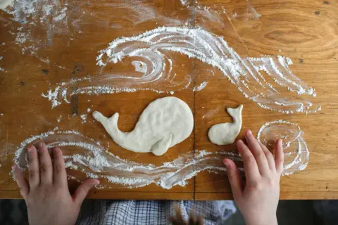 Kinderhände gestalten Salzteig-Ideen: zwei Wale schwimmen in Meereswellen aus Mehl auf einem Holztisch.