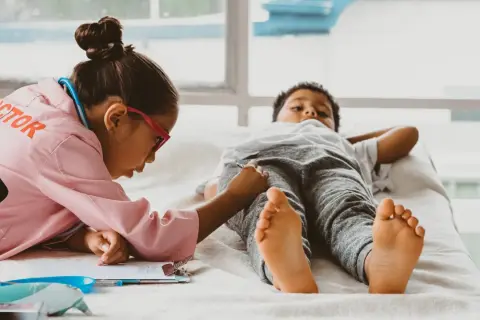 Kinder spielen Doktorspiele im Kindergarten. Ein Mädchen in Arztkittel untersucht einen Jungen, der auf einer Liege liegt.