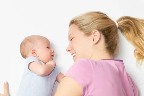 Ein lächelnder baby 2 wochen alt schaut seine Mutter an. Sie liegt neben ihm und lächelt zurück.
