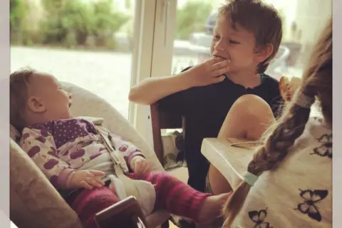 Drei Kinder am Tisch: Ein Baby lacht, ein Junge isst, ein drittes Kind mit Zopf ist im Vordergrund.