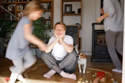 Chaos im Wohnzimmer: Kinder rennen herum, ein Hund schaut neugierig, und die mama steht kopf. Sie sitzt auf dem Boden, die Augen geschlossen, während das Leben um sie herum tobt.