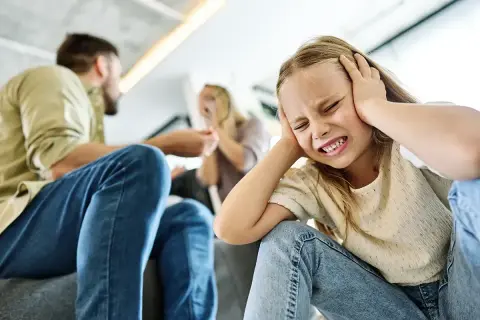 Ein Kind hält sich die Ohren zu, während Eltern im Hintergrund streiten. Das aggressive Verhalten der Erwachsenen belastet das Kind sichtlich.