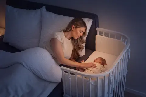 Mutter beruhigt ihr Baby im Beistellbett, um die Schlafregression zu überstehen. Sanftes Licht vom Nachttisch.