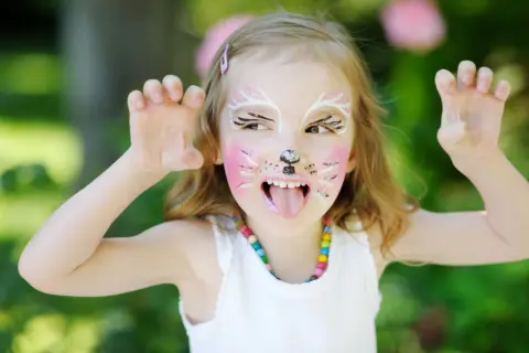 Ein Kind mit süßem Katzen-Make-up, das mit den Pfoten nach oben zeigt. Das Kind hat rosa Wangen, Schnurrhaare und eine schwarze Nase.