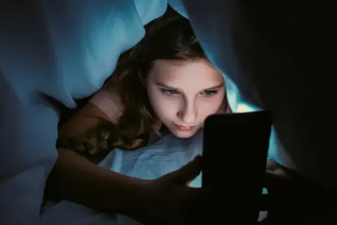 Ein Mädchen liegt im Bett unter der Decke und schaut auf ihr Handy. Sie wirkt vertieft, fast wie faule Kinder, die ihre Zeit mit digitalen Medien verbringen.