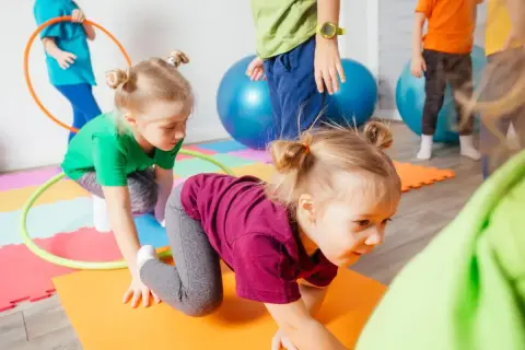 Kinder haben Spaß bei Bewegungsspielen im Kindergarten. Sie krabbeln auf bunten Matten, umgeben von Gymnastikbällen und Reifen.