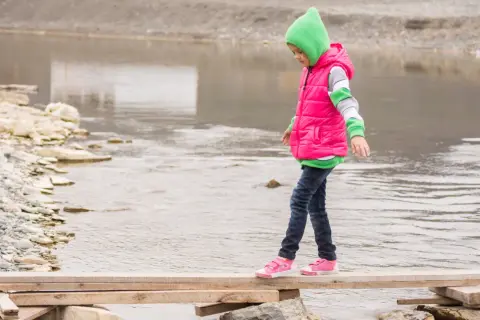 Ein Kind balanciert über ein provisorisches Holzbrett über einem Fluss. Das zeigt, was bedeutet selbstwirksamkeit: Mut, Vertrauen in die eigenen Fähigkeiten und das Überwinden von Hindernissen.