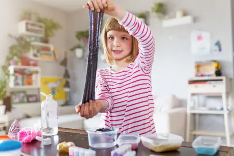 Ein Mädchen hält glitzernden, dunklen Schleim hoch, den sie gerade selber gemacht hat. Auf dem Tisch liegen Zutaten für weitere Schleim-Kreationen.