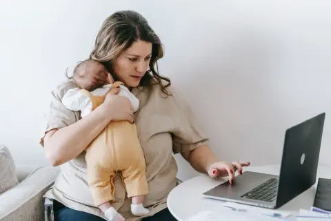 Alleinerziehend und im **burnout**: Eine Frau hält ihr Baby und arbeitet am Laptop.
