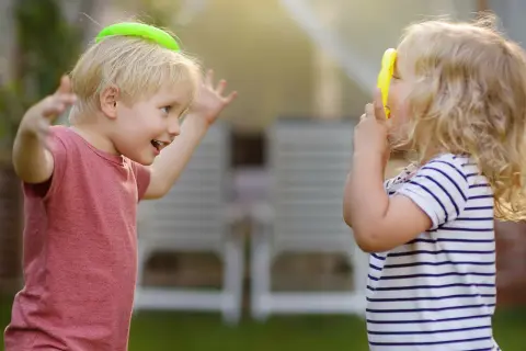 Zwei Kinder spielen im Garten. Der Junge mit einem grünen Ring auf dem Kopf lacht, das Mädchen hält ein gelbes Spielzeug. Ein Beispiel für gelebte sozialkompetenz definition.