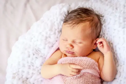 Ein schlafendes Baby, 9 Wochen alt, eingewickelt in eine rosafarbene Decke, liegt auf einer weißen, flauschigen Unterlage.