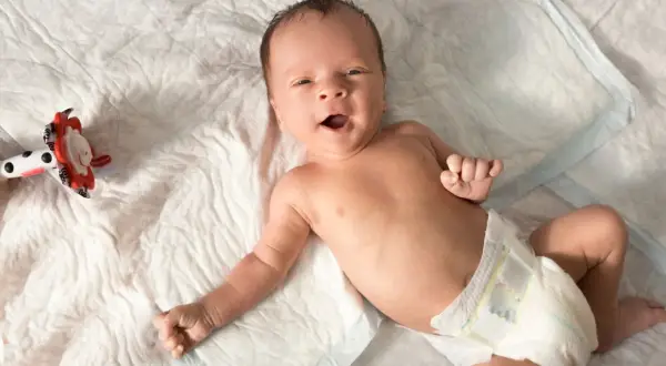 Ein fröhliches Baby, etwa 6 Wochen alt, liegt auf einer weißen Unterlage und blickt neugierig in die Kamera. Neben ihm liegt ein buntes Spielzeug.